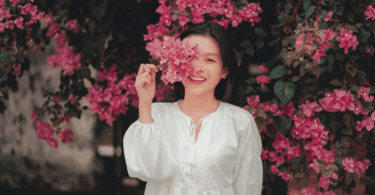 Mulher sorridente segurando flores cor-de-rosa diante do rosto, em meio a um jardim com muitas flores ao fundo.