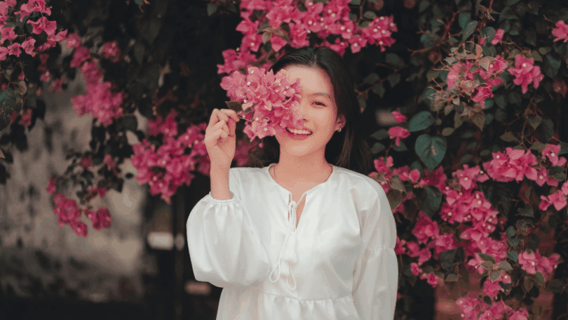 Mulher sorridente segurando flores cor-de-rosa diante do rosto, em meio a um jardim com muitas flores ao fundo.