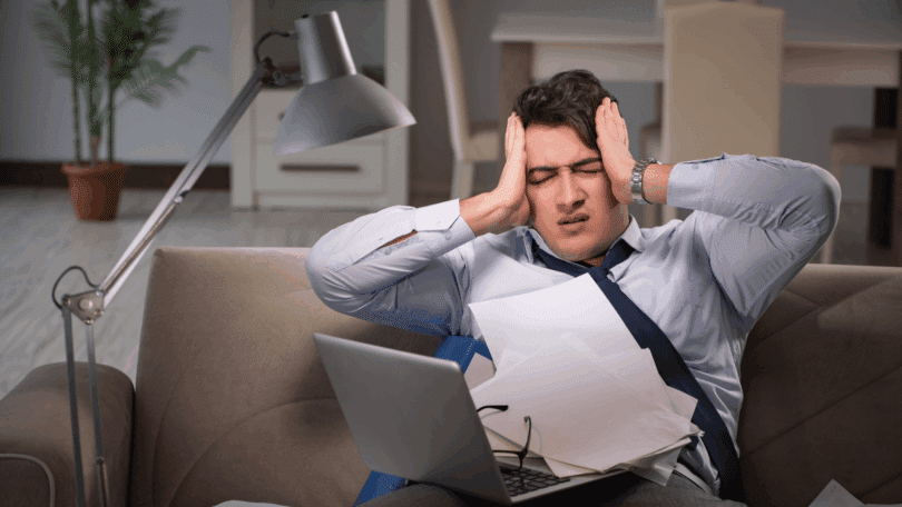 Homem sentado no sofá, com expressão de estresse, segurando a cabeça enquanto lida com papéis e um notebook.
