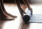 Pessoa desenrolando um tapete de yoga no chão de madeira, com os pés descalços ao lado.