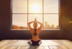 Imagem de uma mulher usando roupa de ginástica, praticando yoga dentro de uma sala com uma janela grande. Ela está olhando para o Sol.