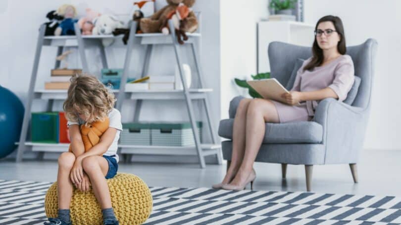 Imagem de uma criança retraída sentada em um pufe em uma sala de brinquedos enquanto a sua mãe está sentada em uma poltrona trabalhando, simbolizando uma mãe narcisista.