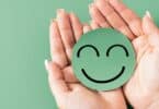 Imagem de fundo verde e sobre ela, as mãos de uma mulher segurando um emoji sorrindo, representando o bem-estar emocional.