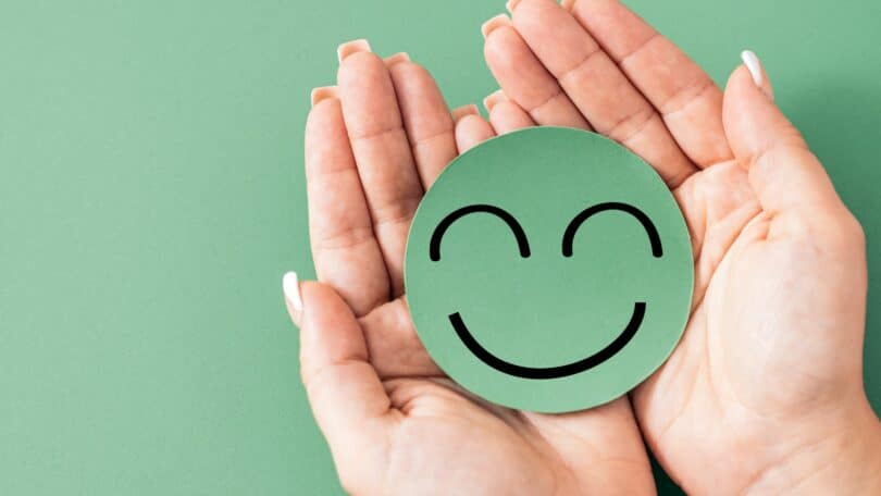 Imagem de fundo verde e sobre ela, as mãos de uma mulher segurando um emoji sorrindo, representando o bem-estar emocional.