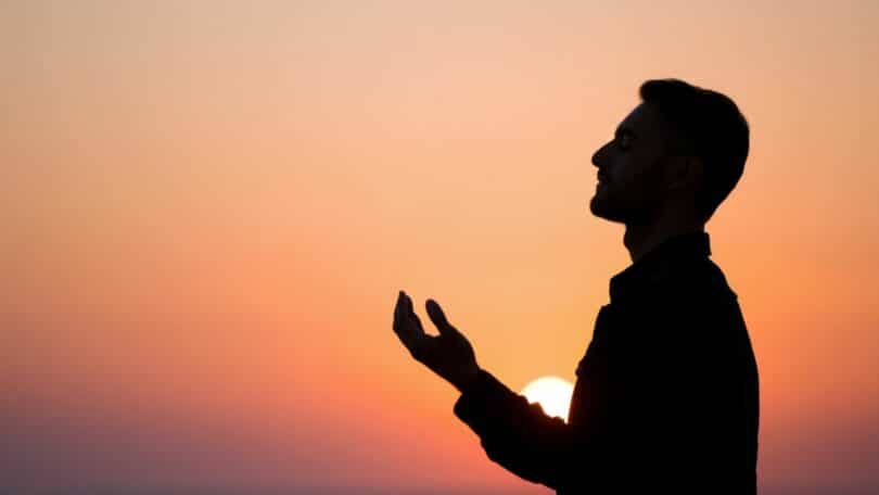 Imagem da silhueta de um homem com as mãos erguidas em sinal de oração. Ao fundo, um lindo pôr do Sol.