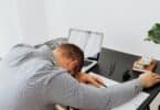 Imagem de um homem debruçado em sua mesa de trabalho, simbolizado o conceito da cultura da hiperprodutividade. Ele está exausto e muito cansado.