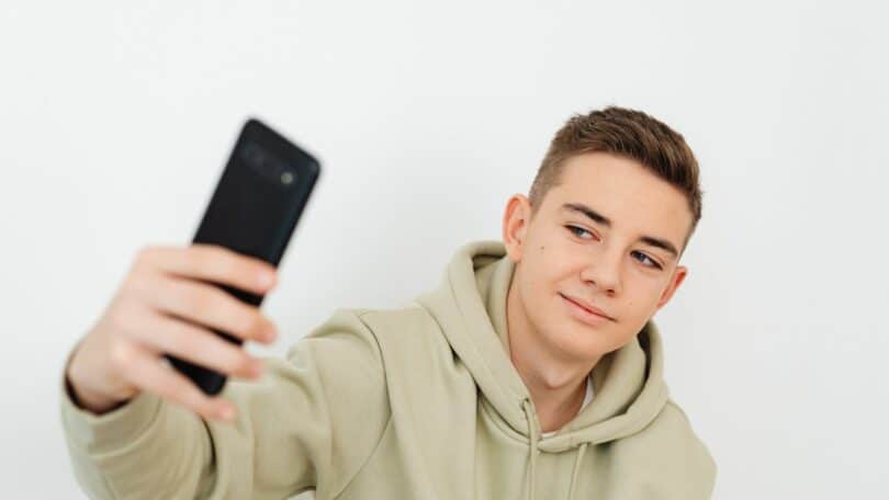 Imagem de um jovem da geração Z, usando um moletom verde claro e fazendo uma selfie com o celular.
