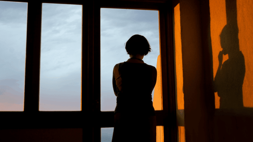 Mulher em silhueta observa pela janela ao entardecer, com sua sombra projetada na parede.