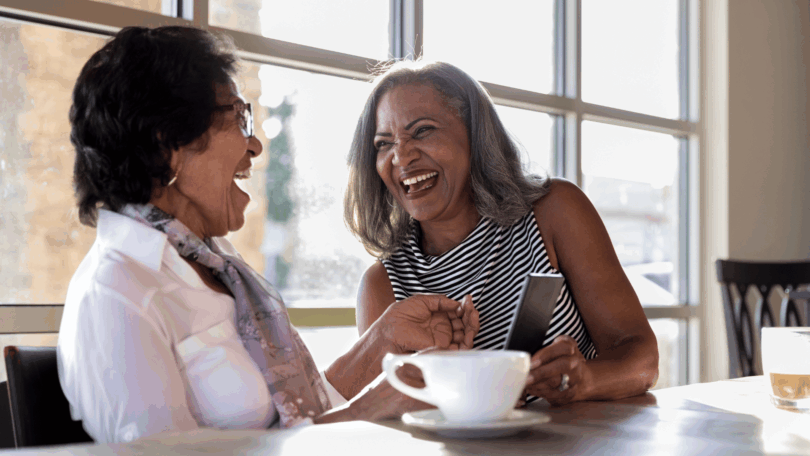 Duas mulheres maduras rindo juntas à mesa de café, conversando de forma descontraída.