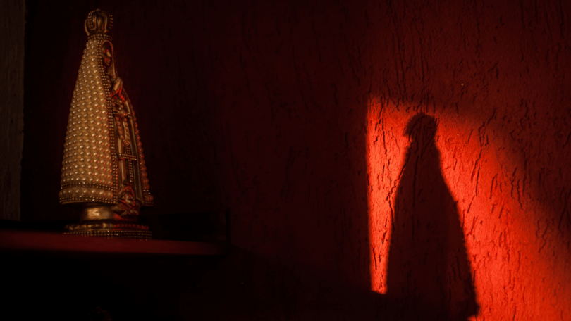 Estátua de Nossa Senhora Aparecida iluminada por luz vermelha, projetando sombra na parede.
