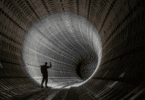 Silhueta de pessoa em um grande túnel metálico curvo, iluminado ao fundo, sugerindo exploração ou desconhecido.