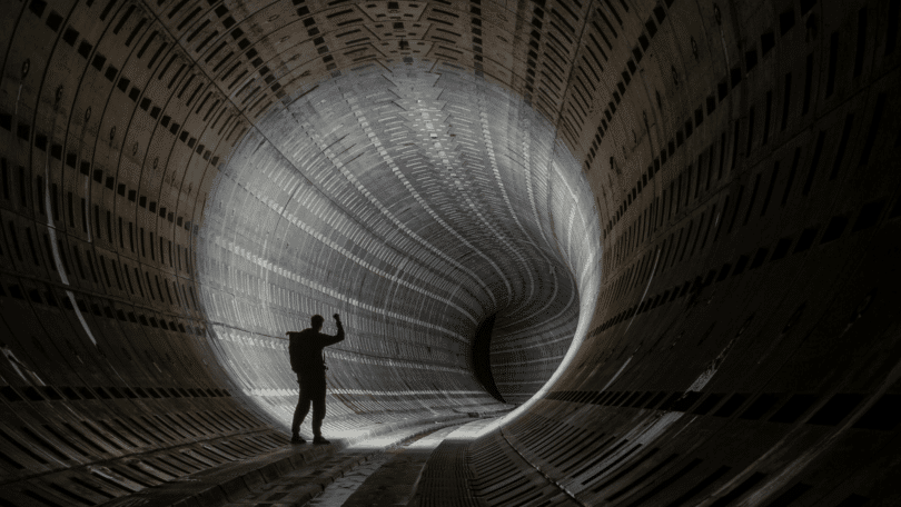 Silhueta de pessoa em um grande túnel metálico curvo, iluminado ao fundo, sugerindo exploração ou desconhecido.