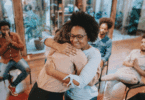 Duas mulheres se abraçam em grupo de apoio, enquanto outras pessoas observam em um ambiente acolhedor.