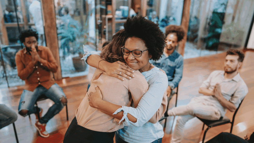 Duas mulheres se abraçam em grupo de apoio, enquanto outras pessoas observam em um ambiente acolhedor.