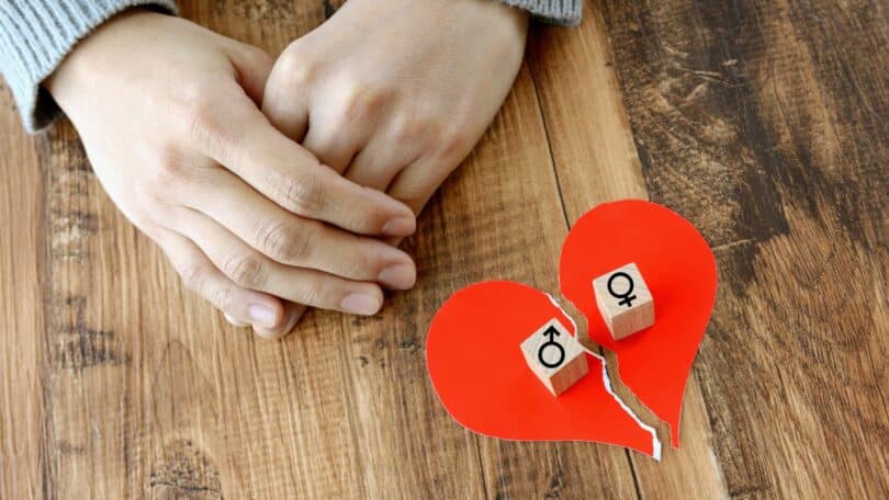 Imagem das mãos de uma mulher apoiada em uma mesa de madeira. Ao lado, um coração feito de papel, cortado ao meio, simbolizando separação entre um casal.