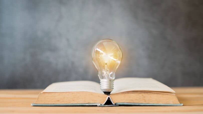 Imagem de um livro aberto sobre uma mesa de madeira e sobre ele, uma lâmpada acesa, simbolizando a sabedoria, o questionamento, a busca pela informação e conhecimento.