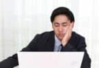 Imagem de um homem de terno, sentado à frente do seu computador em sua mesa de trabalho. Seu semblante é triste e cansado, simbolizando não gostar do seu trabalho.