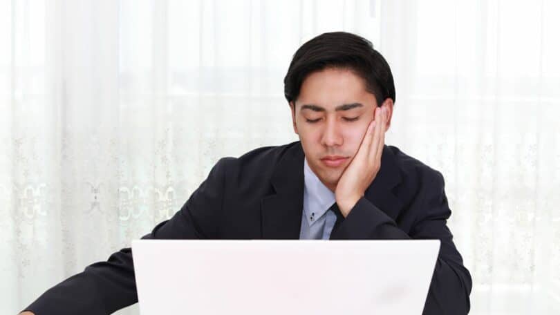 Imagem de um homem de terno, sentado à frente do seu computador em sua mesa de trabalho. Seu semblante é triste e cansado, simbolizando não gostar do seu trabalho.
