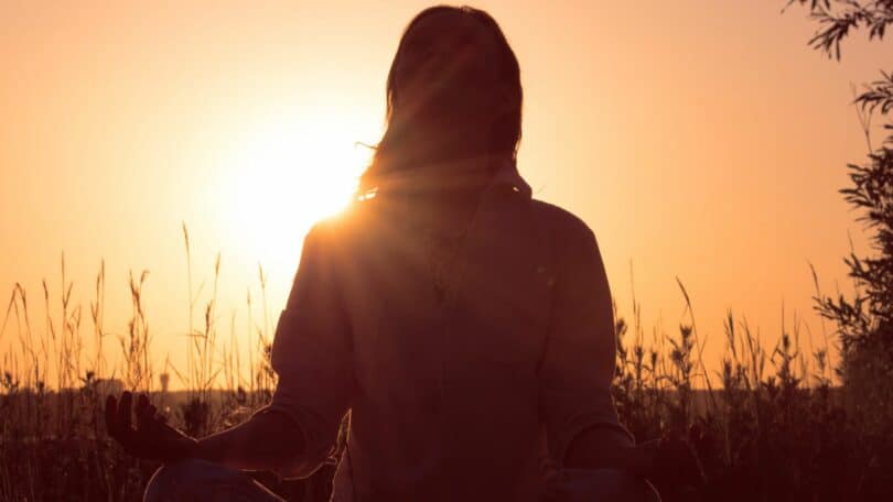 Imagem de uma mulher meditando em um campo de trigo e ao fundo um lindo pôr do sol. A foto traz o conceito do silêncio que cura e de uma mente desacelerada.