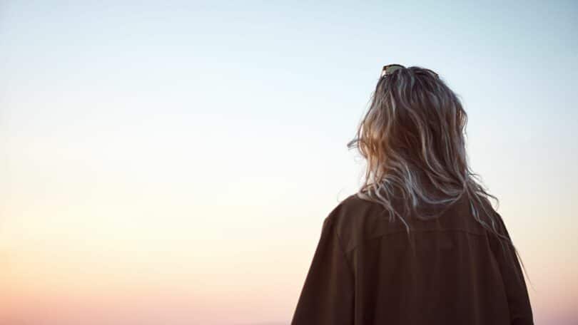 Imagem de uma pessoa de costas, olhando para o horizonte e um lindo pôr do sol. O ambiente é calmo e reflete a natureza.