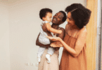 Duas mulheres sorrindo enquanto seguram e interagem com um bebê em ambiente interno iluminado.