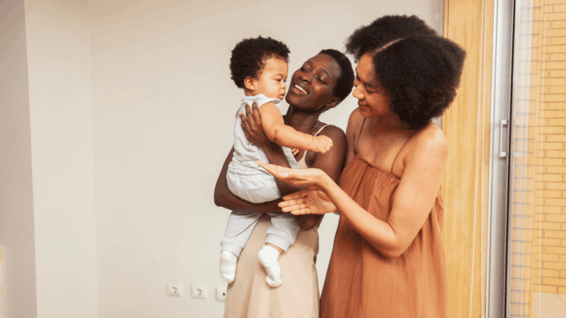 Duas mulheres sorrindo enquanto seguram e interagem com um bebê em ambiente interno iluminado.