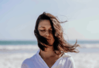 Mulher de olhos fechados na praia com o cabelo sendo levado pelo vento.