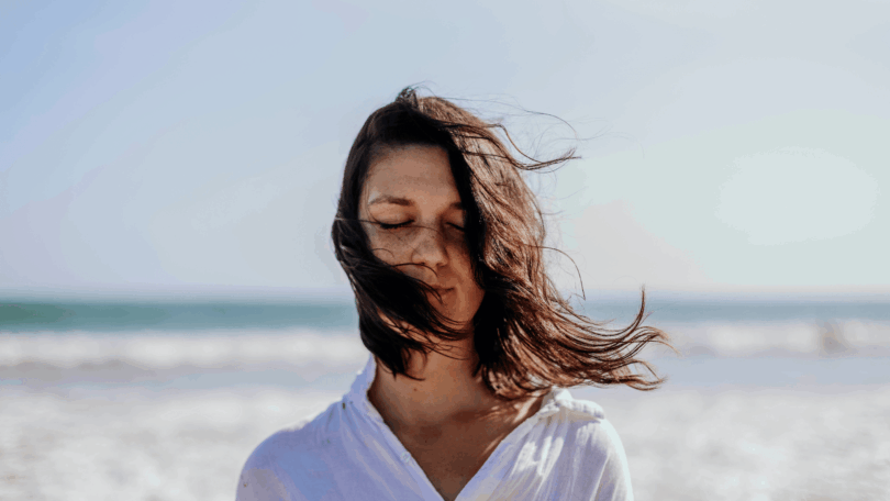 Mulher de olhos fechados na praia com o cabelo sendo levado pelo vento.