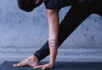 Pessoa pratica uma postura de yoga sobre um tapete, com o braço apoiado no chão e tatuagem “ahimsa” em destaque.