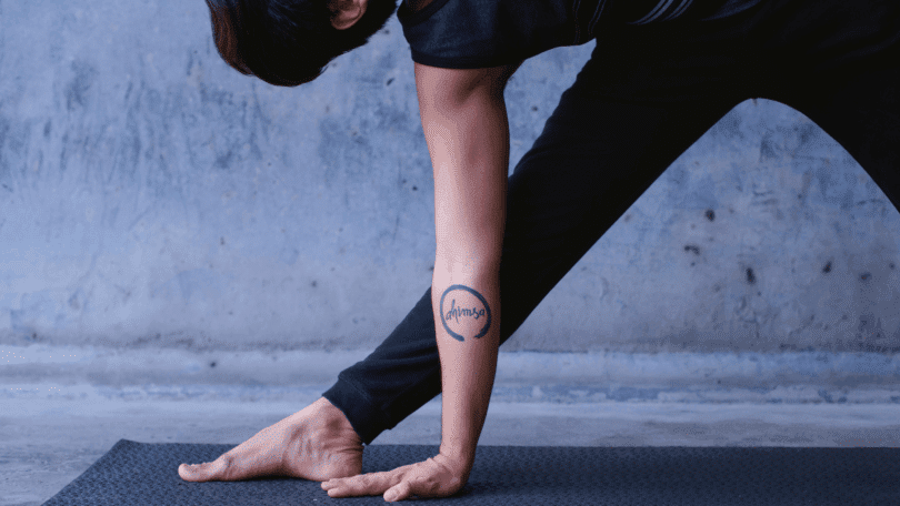 Pessoa pratica uma postura de yoga sobre um tapete, com o braço apoiado no chão e tatuagem “ahimsa” em destaque.