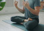 Pessoa sentada em posição de meditação sobre um tapete, com as mãos erguidas em gesto de concentração e equilíbrio.
