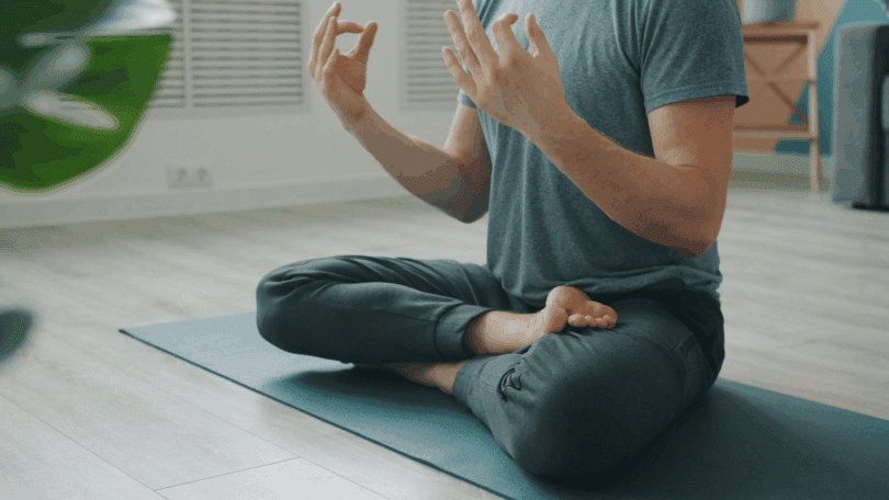 Pessoa sentada em posição de meditação sobre um tapete, com as mãos erguidas em gesto de concentração e equilíbrio.