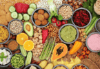 Variedade de alimentos naturais e vegetais organizados em mesa, com grãos, frutas, legumes e preparações saudáveis coloridas.