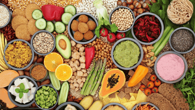 Variedade de alimentos naturais e vegetais organizados em mesa, com grãos, frutas, legumes e preparações saudáveis coloridas.