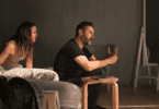 Casal em clima de tensão emocional conversa na sala, enquanto o homem gesticula seriamente e a mulher observa aflita ao lado do sofá.