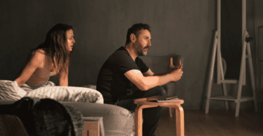 Casal em clima de tensão emocional conversa na sala, enquanto o homem gesticula seriamente e a mulher observa aflita ao lado do sofá.