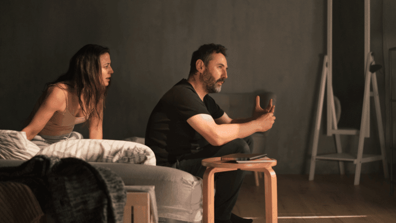 Casal em clima de tensão emocional conversa na sala, enquanto o homem gesticula seriamente e a mulher observa aflita ao lado do sofá.