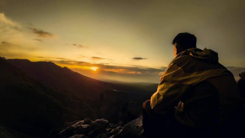 Imagem de um homem sentado em uma pedra no alto de uma montanha, contemplando o universo e o lindo pôr do Sol.