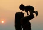 Imagem da silhueta de uma mãe segurando o seu filho no alto. Ao fundo, um lindo pôr do Sol. A imagem simboliza a mulher e os desafios de ser mãe.