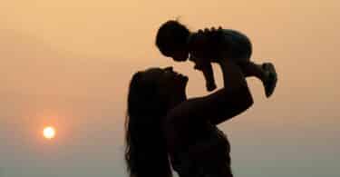Imagem da silhueta de uma mãe segurando o seu filho no alto. Ao fundo, um lindo pôr do Sol. A imagem simboliza a mulher e os desafios de ser mãe.