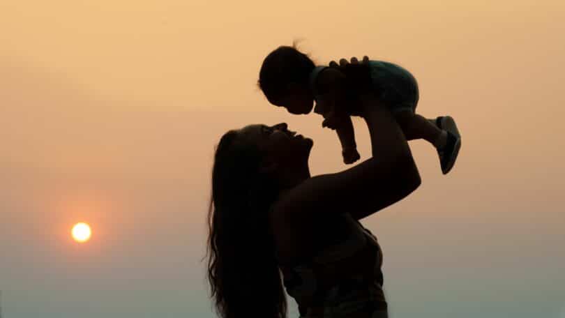 Imagem da silhueta de uma mãe segurando o seu filho no alto. Ao fundo, um lindo pôr do Sol. A imagem simboliza a mulher e os desafios de ser mãe.