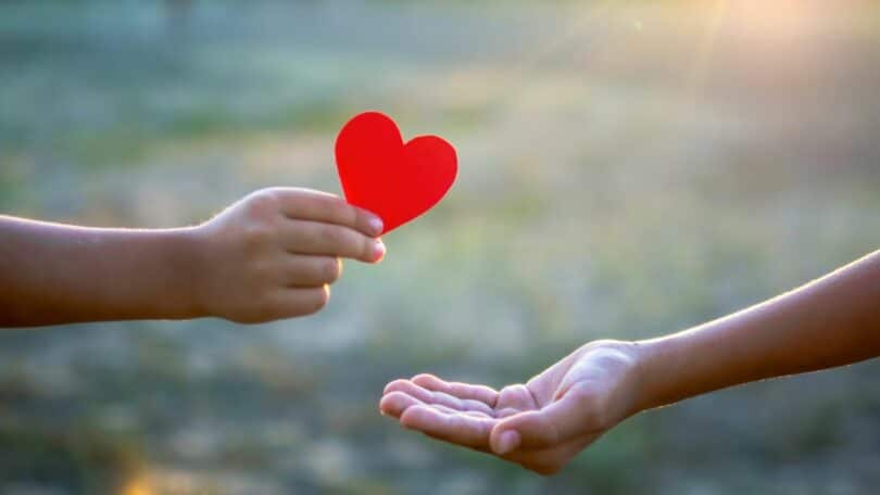 Imagem de duas mãos - de uma criança e sua mãe. A criança está está entregando um coração vermelho de papel para a mãe, simbolizando o conceito de ser mãe.