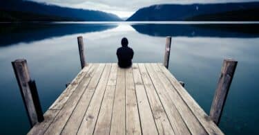 Imagem de uma pessoa sentada em um pier de madeira, olhando para um lago. A foto traz um conceito existencial, introspectivo e sofisticado, transmitindo solitude e autonomia.