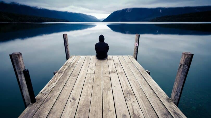 Imagem de uma pessoa sentada em um pier de madeira, olhando para um lago. A foto traz um conceito existencial, introspectivo e sofisticado, transmitindo solitude e autonomia.