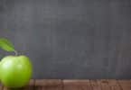 Imagem de um quadro negro ao fundo de uma sala de aula e em destaque uma maçã na cor verde, sobre a mesa do professor, simbolizando a educação.