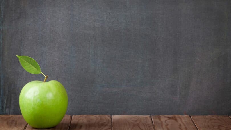 Imagem de um quadro negro ao fundo de uma sala de aula e em destaque uma maçã na cor verde, sobre a mesa do professor, simbolizando a educação.
