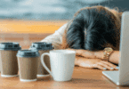 Mulher descansando a cabeça sobre a mesa ao lado de um notebook e vários copos de café, sugerindo cansaço, exaustão ou sobrecarga de trabalho.