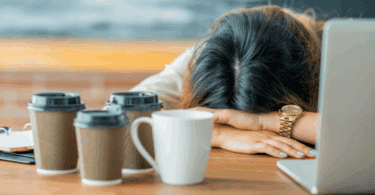 Mulher descansando a cabeça sobre a mesa ao lado de um notebook e vários copos de café, sugerindo cansaço, exaustão ou sobrecarga de trabalho.