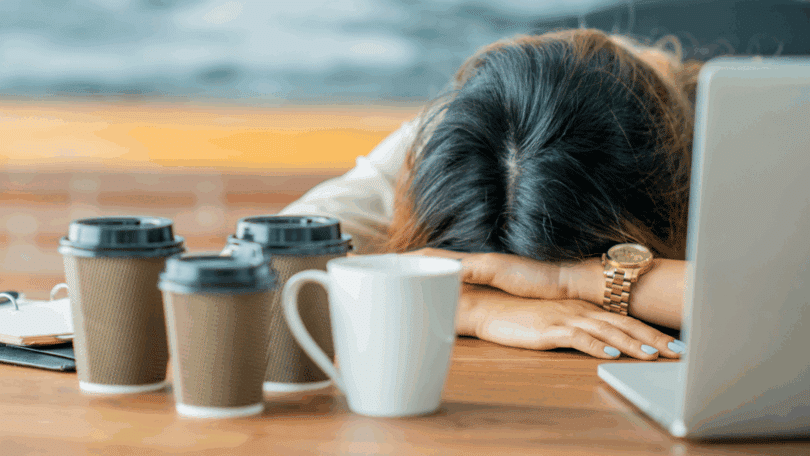 Mulher descansando a cabeça sobre a mesa ao lado de um notebook e vários copos de café, sugerindo cansaço, exaustão ou sobrecarga de trabalho.