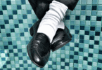 Pernas cruzadas com sapatos pretos e meias brancas diante de um fundo de azulejos azuis, em composição de estilo retrô e urbano. Os sapatos lembram o estilo do Michael Jackson.